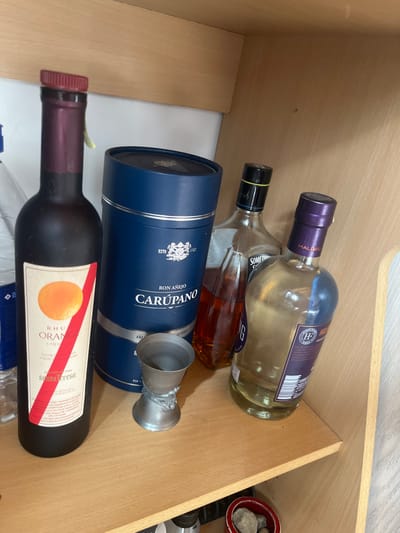 The image displays an interior scene within a wooden storage unit or cabinet, located in Ushuaia, Argentina. A collection of alcoholic beverages and a single measuring jigger are arranged on a light-colored wooden shelf. On the far left, a clear plastic bottle is partially visible. Next to it stands a dark brown glass bottle of Santa Teresa Rhum Orange Liqueur, featuring an orange circle graphic and a red diagonal stripe on its label. Adjacent to the liqueur bottle is a dark blue cylindrical presentation box for Ron Añejo Carúpano, marked with the brand name, "RON AÑEJO," and "ESTD 1767." In front of the Carúpano box, a metallic double jigger, with a detailed decorative base, rests on the shelf. To the right, a clear glass bottle filled with an amber-colored spirit is present, featuring a black cap with a yellow band. Beside it is another clear glass bottle containing a pale yellow liquid, identifiable by a purple neck wrap labeled "MALBEC" and an "HP" logo on its main label. The background behind the shelf is a plain white surface. A small, red, circular container holding several grey rocks is partially visible on a lower shelf in the bottom right corner of the frame.