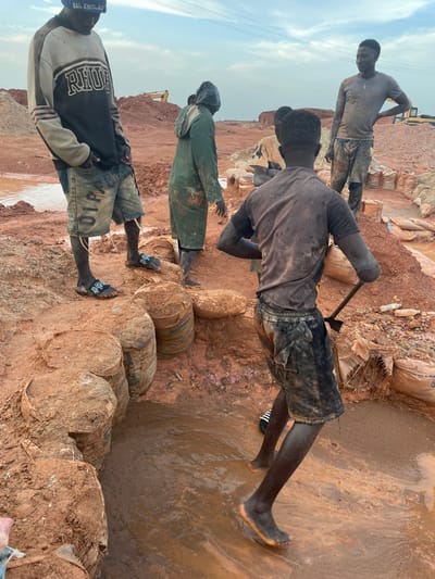 The image depicts a group of young Black men working in a muddy, open-air excavation site under a cloudy sky, suggesting it's daytime. The ground is composed of reddish-brown soil, with visible puddles of water indicating recent or ongoing wet conditions. Several large, roughly cylindrical objects wrapped in burlap sacks are stacked on the ground, possibly containing raw materials or waste.

One man, positioned prominently in the foreground and facing away from the camera, is wearing dark shorts and a gray, dirty t-shirt. He is actively using a shovel, digging into the muddy ground. Another man, slightly further back, wears a green long-sleeved garment and a head covering. In the background, two other men are visible, one of whom is standing and looking towards the left, wearing a t-shirt and shorts. The final man, partially visible at the bottom left, is wearing a black and gray sweatshirt with white lettering and a blue beanie. He is also wearing shorts and sandals.

The overall scene suggests manual labor in a challenging environment, with the men appearing focused on their tasks. There are no clear indicators of emotion, but the exertion of digging in the mud is implied. In the far background, a yellow excavator is partially visible, hinting at larger-scale operations at the site. Some of the burlap sacks have visible text, including "SOLO" and what appears to be "PREMIUM CEMENT," suggesting they might be related to construction materials or waste from such operations. The location is identified as Rayfield, Nigeria.