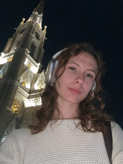 A woman with curly brown hair and wearing white headphones and a light-colored sweater is taking a selfie at night. Behind her is the illuminated facade of the Name of Mary Church, a Roman Catholic church located in Novi Sad, Serbia. The church is characterized by its Gothic Revival architecture, including a tall spire, pointed arches, and intricate stonework. The sky is dark.