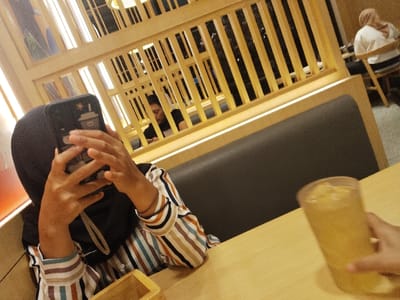 The image shows a young woman sitting at a table in a restaurant in Telukjambe, Indonesia (latitude: -6.301, longitude: 107.278). She is wearing a striped shirt and a black headscarf. The woman is holding a smartphone in front of her face. A glass of iced tea is on the table next to the woman's right hand.  A wooden slatted partition separates the table from another seating area. A second person is partially visible behind the partition, seated with their back turned towards the camera. In the background, a person is sitting in a chair, wearing a white shirt and a brown headscarf. The wooden table has a glossy surface and is illuminated by overhead lighting. The environment is clean and well-lit.
