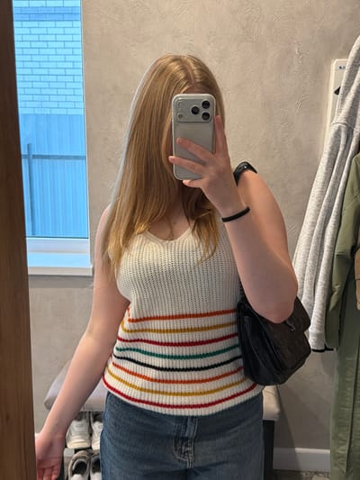 A person stands in front of a mirror, holding a smartphone to take a selfie. The person has long blonde hair and wears a white knitted top with vertical colored stripes. The person also wears blue jeans. A black backpack rests on the person's shoulder. In the background, there is a rack of clothing, a textured wall, a window, and a wooden frame around the mirror. The location appears to be indoors. The city of Rossiyskiy, Russia, cannot be determined from the image.