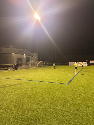 The image displays an artificial turf soccer field at night in the city of Newton, United States. The scene is illuminated by multiple artificial light sources, most prominently a tall stadium light pole in the upper left, casting significant glare across the upper portion of the frame. The sky is dark.
The field itself features green synthetic turf with distinct blue demarcation lines. Along the left edge of the field, a white metal soccer goal with a net is visible, situated near a two-story light-colored building. The building has windows and rooftop units. A yellow line runs horizontally across the field edge in front of the building.
On the field, four individuals are present. Three males are positioned on the artificial turf in the mid-ground and foreground, all wearing bright yellow-green short-sleeved shirts. The male closest to the viewer, in the center-right foreground, also wears dark shorts and stands with hands on his hips. A second male, further back and to his left, also wears dark shorts. A third male, further back and to his right, wears red shorts. These three individuals are positioned within the field's playing area. A fourth individual, dressed in darker clothing, stands near the soccer goal on the left side of the field, closer to the building. In the background, beyond the field, trees and dark fencing are discernible.