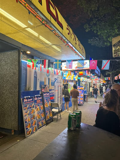The image depicts a lively street scene at night, likely in Vancouver, Canada, judging by the prevalence of international flags and the general urban environment suggestive of a multicultural city. In the foreground, a dark, possibly wooden, table is occupied by three green cans of "Tempo" soda. To the right, a person in a black top is partially visible, facing away from the camera.

The main focus of the image is a row of Mediterranean-themed food stalls or restaurants lining a sidewalk. These establishments are adorned with numerous international flags strung across their awnings, creating a festive atmosphere. Prominently displayed are large menu boards with pictures of food items and prices, such as "Chicken Shawarma Pita" for $15.99 and "Beef & Lamb Donair Plate" for $18.99. The architecture suggests a compact, perhaps older, urban setting.

Several individuals are seen walking along the sidewalk, engaged in conversation or browsing the menus. The lighting is a mix of string lights, illuminated signs, and the ambient glow of the street. Trees with dark foliage are visible in the background, suggesting an outdoor, possibly pedestrian-friendly, area. The overall impression is one of a bustling, diverse, and vibrant dining district at night.