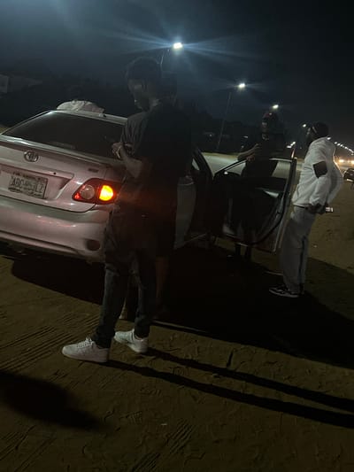 The image shows a night scene in what appears to be Galadimawa, Nigeria, based on the license plate on the back of a silver Toyota car that reads "Abuja." A young man wearing a dark shirt and pants with white sneakers is standing near the open back door of the car. There's another individual in a white outfit standing behind the car, partially obscured. Street lights illuminate the area. The ground appears sandy or uneven. The action seems focused around the car, with people potentially interacting or accessing it.