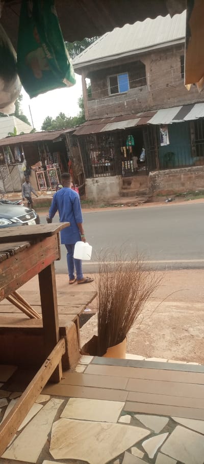 The image captures an urban street scene in Ogidi, Nigeria, viewed from beneath an overhead structure, likely a shop canopy.
In the immediate foreground, a section of a reddish-brown wooden market stall is visible on the left. Suspended from an unseen overhead structure, a green plastic bag displaying a "MAYO" brand label and a lighter-colored bag are visible. To the right, a bundle of dry, thin brown plant stalks, consistent with broom bristles, stands upright in a cylindrical brown container. The immediate ground features light-colored, irregularly shaped and rectangular floor tiles.
A paved asphalt road extends horizontally across the midground. A male figure, viewed from behind, is walking leftward on the sidewalk adjacent to the road. He wears a blue tunic and matching trousers with brown open-toed sandals, and carries a translucent white plastic jerrycan with a handle in his right hand. A dark-colored Lexus sedan is partially visible on the road's left side. Further left on the sidewalk, a second male figure, dressed in a plaid shirt, walks away from the viewer.
Across the road, a series of single-story commercial structures with corrugated metal roofs, some showing rust, are present. These shops display various goods, including packaged items and clothing, often behind metal grilles. Behind and above these single-story structures, a two-story building with a light-colored corrugated metal roof is visible. Its facade is a reddish-brown, and it features a window with a blue-tinted pane in a white frame. Green foliage is discernible behind the buildings. The sky above is bright, suggesting daylight.