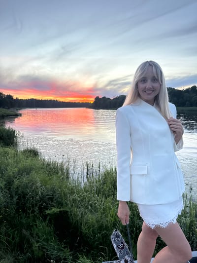 The image shows a young woman standing outdoors near a body of water during what appears to be sunset. She is smiling and looking towards the camera. She is wearing a white blazer over a white lace-trimmed outfit. Her blonde hair is framing her face. She is holding a small, dark, patterned bag by a strap in her right hand. The background features a body of water that reflects the vibrant colors of the sunset, with hues of orange, pink, and yellow in the sky. Green foliage is visible on the right side of the image and along the shoreline. The time of day is evident from the dramatic sunset colors and the soft lighting. The overall mood seems peaceful and happy. The location is identified as Carnikava, Latvia. There is no visible text in the image.
