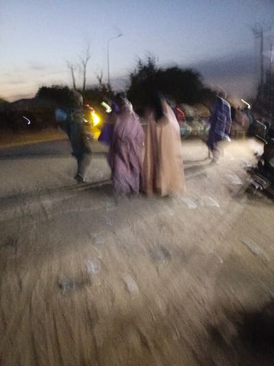A blurry, low-lit image shows several figures standing or walking on a rough surface. Three figures in the center wear long garments in shades of brown and purple. A fourth figure to the left is clothed in darker colors. A street lamp is visible in the background. The overall impression is one of movement and indistinct forms due to the blur.
