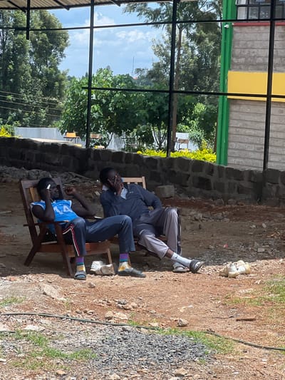 The scene depicts two adult males seated in wooden chairs within an outdoor, semi-enclosed common area in Nyeri, Kenya. The ground surface is predominantly reddish-brown dirt and gravel with scattered rocks. An overhead structure composed of black metal beams, suggesting a roof or canopy framework, is visible.

The male on the left, with a dark complexion, wears a blue sleeveless vest with white numerals, dark trousers featuring a side stripe, multi-colored striped socks, and dark sandals. He holds a white earpiece to his left ear, maintaining a relaxed posture. The male on the right, also with a dark complexion, wears a dark grey long-sleeved polo shirt, light grey trousers, white socks, and dark sandals. He is seated in a relaxed position with his right hand near his mouth. No direct interaction between the two individuals is observed.

On the ground adjacent to the right male, three pairs of light-colored athletic shoes are present, and a single white sneaker lies near the left male's feet.

A low wall constructed of dark grey blocks or stones runs across the midground. Beyond this wall, dense green tree foliage dominates the background. To the far right, a multi-story building is visible, featuring a light grey block facade with a prominent green vertical stripe and a yellow horizontal stripe. In the distant background, a communication tower stands on a hill against a partly cloudy sky. Several additional empty wooden chairs are positioned further back, beyond the low wall.