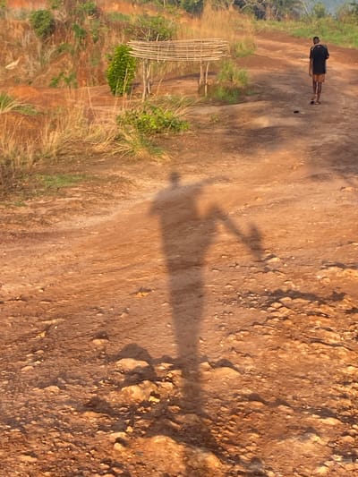 The image shows a person walking on a dirt path in a rural area. A shadow suggests a person holding a fishing rod in the middle of the path. The area is dry and grassy, with some scrub vegetation and rocks. To the left of the frame, what appears to be a bamboo structure is visible. Based on the location provided, this scene likely represents a rural area surrounding the city of Amoso, Nigeria. The light suggests either early morning or late afternoon. The person is walking to the right of the frame and wearing dark clothing.