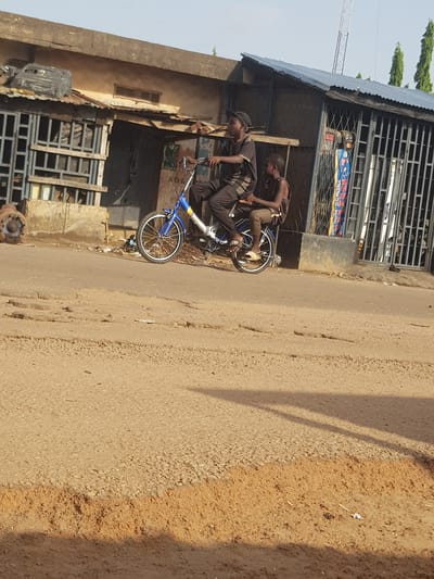 Two individuals are depicted on a bicycle on a dirt road in Kufena, Nigeria. The person in the front, wearing a black and grey striped t-shirt and black pants, is operating the bicycle. Seated behind them, facing forward, is another individual wearing a dark sleeveless shirt and light brown pants. The bicycle has a blue frame and white tires with yellow rims. In the background, a building with a corrugated metal roof and bars on its windows is visible. The environment appears to be an outdoor, possibly urban or semi-urban setting, characterized by the unpaved road and the utilitarian nature of the building.