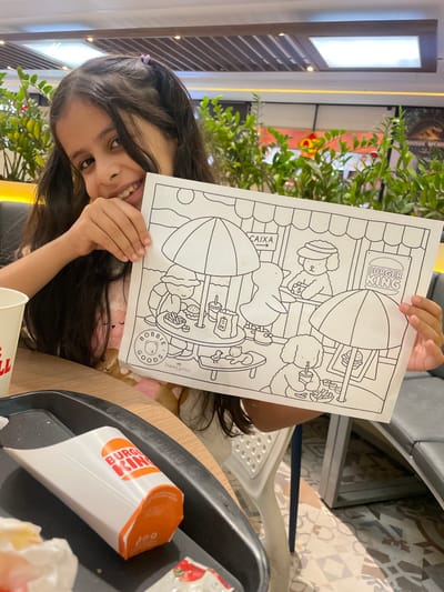 The image displays a young girl, estimated to be between 6 and 10 years old, seated at a light-colored table in a Burger King restaurant, likely situated within a food court in Anápolis, Brazil. She has long, dark hair, styled with a small purple clip on her left side, and is smiling towards the camera.

The girl holds a black-and-white coloring page prominently in the center of the frame. The page depicts anthropomorphic animal characters in an outdoor cafe setting with umbrellas. Visible text on the coloring page includes "CAIXA" (Portuguese for "Cashier" or "Checkout") with an arrow, a sign reading "BURGER KING," and branding for "BOBBIE GOODS" (with a bear logo) and "Faber-Castell."

On a black fast-food tray in the foreground, an orange and white Burger King french fry container is present, alongside a white paper cup and a small, red-and-white packet, likely containing condiments.

The background shows lush green potted plants, a common feature in food court environments. Overhead lighting fixtures, including rectangular recessed lights, are visible on the ceiling. Partially visible behind the plants on the right is a poster or display featuring "JURASSIC WORLD" branding. The floor beneath the table and chairs is covered in patterned tiles with geometric designs in various shades. Seating visible includes a white chair with a perforated back behind the girl and a gray padded bench seat to the right.