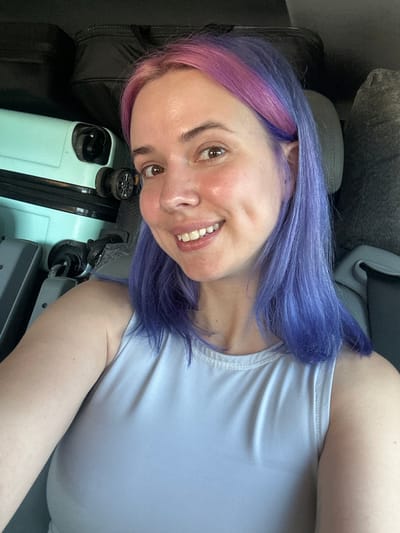 A selfie captures a woman with dyed purple and pink hair, smiling, positioned near a light blue suitcase. The background consists of a vehicle interior including what appears to be car seats. The image does not provide any information that can be used to pinpoint the exact location in the city of Varazze, Italy.