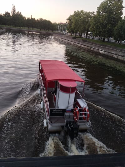 An aerial view captures a red-canopied pontoon boat moving at speed through the Pregolya River in Kaliningrad, Russia. The boat is leaving a strong wake. On the boat, life preservers are visible. The riverbank is lined with concrete structures and tall, leafy trees. In the distance, buildings and infrastructure can be seen. The sky is a pale, dusky hue, indicating either early morning or late evening.