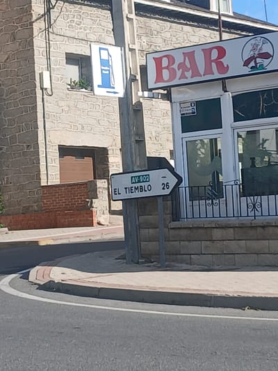 The image shows a street scene in Navaluenga, Spain. In the foreground, there is a paved road with a white line marking a curve. A concrete curb with a pinkish-tan edge separates the road from a sidewalk. Behind the curb and sidewalk, a gray pole stands vertically. Attached to this pole is a white directional sign with a black border. The sign has a blue gas pump symbol on the left, pointing to the right with an arrow. To the right of the gas pump symbol, the text "AV-902 EL TIEMBLO 26" is written in black.

To the right of the sign and pole, a building with a stone facade is visible. A white and red sign with the word "BAR" in red, stylized lettering is mounted above the building's entrance. To the right of the "BAR" sign is a circular logo with a cartoon character. The building has large windows with dark frames and what appears to be a black metal railing in front of them.

Further to the left, another building with a stone facade is visible. This building has a window with plants on the sill and a closed brown door or garage-like entrance. A white electrical box is mounted on the wall of this building. A white sign with a blue gas pump icon and an arrow pointing right is also attached to the stone wall. The overall scene appears to be a sunny day.