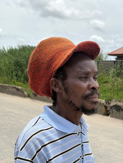 The image presents a close-up, side profile shot of an adult Black man looking upwards. He is wearing a distinctive orange, textured, knitted cap and a light blue polo shirt adorned with vertical black and thin white stripes. The man has a full beard and mustache. His eyes are open, and his gaze is directed towards the upper right of the frame (when upright). The background is an outdoor scene under a cloudy sky, suggesting daytime. Green foliage, likely bushes or trees, is visible behind him to his left, and a paved or concrete surface stretches into the background. A hint of a building or structure can be seen in the distance on the upper right. The man's expression is neutral. No visible text is present. The setting is Igboefon, Nigeria.