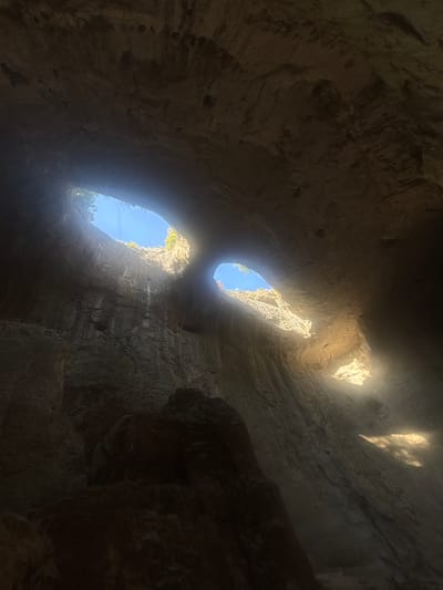 The image captures the Prohodna Cave, also known as the Eyes of God cave, located in Karlukovo, Bulgaria. The cave features two large, eye-shaped openings in its ceiling, through which sunlight streams into the cavern. The rock formations within the cave are rough and textured, with visible striations indicating geological processes. Vegetation can be seen growing around the edges of the openings in the ceiling. The overall impression is one of natural grandeur and a unique geological formation.
