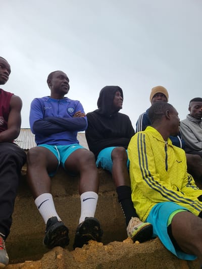 The image, rotated 90 degrees clockwise, captures a low-angle perspective of at least six men resting outdoors against a light, overcast sky in Rafin Bauna, Nigeria. The men are situated on rugged, sandy, and earthy terrain, looking upwards.
In the mid-ground, a man wears a vibrant yellow Adidas jacket with distinctive blue stripes on the sleeves and blue athletic shorts. His light-colored athletic shoes are partially visible against the ground. To his left, another man is seen in a black hooded sweatshirt and blue shorts. Behind them, a man in a grey hooded top with blue stripes and another wearing a brown beanie are also looking up.
In the foreground, on the left side, a man in a blue long-sleeved quarter-zip top, adorned with the Inter Milan football club logo on the chest, has his arms crossed while looking upwards. He is wearing blue athletic shorts, white socks featuring a black Nike swoosh, and dark athletic shoes. Another man, partially visible in the bottom left, is wearing a maroon t-shirt.
The scene suggests a moment of rest or observation, possibly during a sports event or training session, given the athletic attire. The sky is uniformly bright grey, indicating an overcast day. The ground texture is coarse, characteristic of natural, unpaved outdoor areas. No strong emotions are overtly displayed, with expressions generally neutral as they gaze skyward.