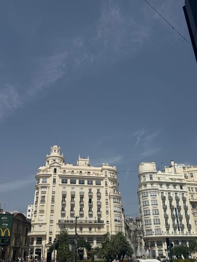 This image captures a scene in Valencia, Spain, specifically the Plaza del Ayuntamiento. Dominating the foreground are two grand, cream-colored buildings with ornate architectural details, including balconies and numerous windows. The building on the left features a distinctive circular turret with a spire. The building on the right is also tall and imposing, with curved facades. In the middle ground, palm trees are visible, and a fountain is spraying water. In the background, more buildings line the street, suggesting a dense urban environment. A few people are visible on the street level, as well as some vehicles. The sky above is a clear, pale blue with sparse, wispy clouds. A green McDonald's sign with yellow arches is partially visible on the left side of the frame. The overall impression is a bustling city square on a bright, sunny day.