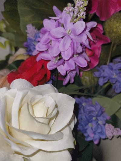 The image displays a close-up, high-angle view of a bouquet of artificial flowers. In the foreground, prominently positioned in the lower-left quadrant, is a white or cream-colored rose with multiple layers of fabric petals, exhibiting slight creasing and wear. Directly behind and to the left of the white rose, a cluster of red fabric flowers is partially visible. The mid-ground and upper-center feature a dense cluster of light purple, possibly lilac-like, fabric flowers with multiple individual florets. To the right of these light purple flowers, a section of a magenta or dark pink flower is visible, along with a blurred, spherical, yellowish-green object in the upper-right corner. Further down and to the right, a cluster of darker blue or purple fabric flowers is present. Green foliage, likely artificial, is visible throughout the background and interspersed among the flower clusters. The lighting suggests an indoor or controlled environment. There are no individuals, actions, or specific interactions depicted. The visual content of the image does not provide any identifiable landmarks, architectural features, or unique environmental cues to ascertain its specific location within the city of Liège, Belgium.