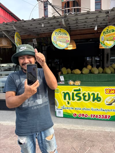 A man stands outdoors, holding a smartphone, in front of a durian stand in Moo 1, Thailand. He is wearing a green Nike hat, a grey-blue t-shirt with "CHALLENGE" printed on it, and ripped blue jeans. The durian stand is displaying numerous durians, along with yellow signs featuring Thai script and images of durians. One sign includes a phone number, 090-1765587. Hanging above are round signs with similar imagery. The environment appears to be a commercial area.