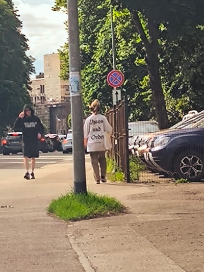 A person wearing a black hoodie and shorts walks on a sidewalk in Riga, Latvia, with a tall building and trees in the background. A second person, seen from behind and wearing a white sweatshirt with text on it and brown pants, walks further down the sidewalk. A dark-colored SUV is parked on the right side of the image, with a fence and foliage behind it. A traffic sign is visible on a pole between the two people. The scene appears to be a street in a city, possibly a residential or commercial area, with cars and pedestrians present. The lighting suggests it is daytime, with scattered clouds in the sky.