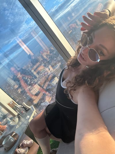 The image shows a woman taking a selfie in a high-rise building overlooking a city. The woman is wearing sunglasses and a black dress with white trim. She has curly brown hair and her hand is raised to her forehead. In the background, the city skyline is visible through a large window. The reflection in the window shows other people and the interior of the room. There is a table with drinks and napkins on it. The image appears to be taken during the daytime, with sunlight streaming in through the window. The specific location in Hanoi, Vietnam cannot be determined from the image alone.