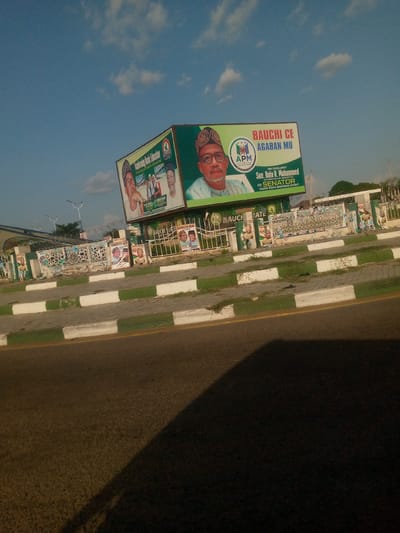 The image captures a roadside scene in Bauchi, Nigeria, dominated by a large, multi-faceted political campaign billboard. The prominent right face of the billboard features a clear portrait of Senator Bala A. Mohammed, wearing glasses and a traditional cap, alongside the logo for the Allied Peoples' Movement (APM) and the slogans "BAUCHI CE AGABAN MU" (Bauchi is our progress). Text below identifies him as "His Excellency Sen. Bala A. Mohammed (KAURAN DAULAN USMANIYYA) FOR SENATOR BAUCHI SOUTH SENATORIAL DISTRICT," confirming the location and political context. The visible left face of the billboard carries the message "Transforming Basic Education, Ensuring no child is left behind," illustrated with various smaller images of individuals.
Below the main billboard, a low concrete wall or fence is densely covered with numerous smaller, often overlapping, political campaign posters, many featuring similar male candidates and including phrases like "BAUCHI STATE KAURARASIYA ACIDA 2023." Barbed wire is strung along the top of sections of this fence. In the foreground, an asphalt road displays a yellow dividing line and a prominent dark shadow stretching across its surface. The road is bordered by a sidewalk featuring a distinct curb painted in alternating white and green blocks, with sparse patches of green grass behind it. The sky above is clear blue with scattered white clouds. In the background, beyond the fence, a building with a blue roof and some trees are visible, along with street light poles. The lighting suggests it is either early morning or late afternoon.