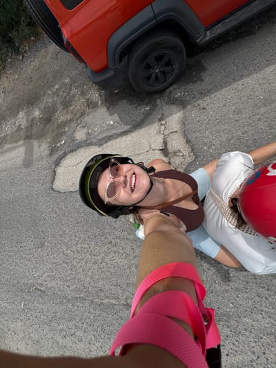 In Baan Bang Rak, Thailand, two individuals, presumably friends, are captured in a selfie-style photograph. The woman in the foreground, wearing a brown tank top and a black helmet with sunglasses, is smiling at the camera. She has a pink strap wrapped around her leg, which appears to be part of a safety harness or backpack. Beside her, another woman is partially visible, wearing a white t-shirt and a red helmet. They are both seated or positioned on some form of transport, likely scooters or a similar vehicle, as suggested by the helmets and their proximity to the road. In the background, a bright red SUV is parked, with its rear wheel and part of its body visible. The ground beneath them is an asphalt road, showing signs of wear and tear with cracks and patches of lighter material. The overall impression is a casual outing or activity, possibly exploring the area in Thailand.