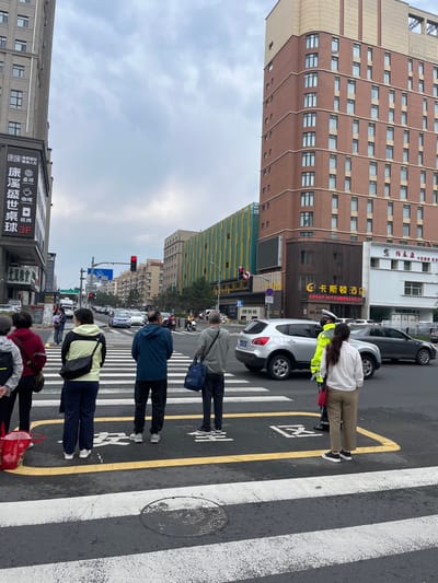 In Changchun City, China, at a street intersection, several pedestrians are waiting to cross the street at a crosswalk. The sky is overcast, and traffic is present. On the left side of the image, a tall building with a sign featuring Chinese characters and the number "3F" is visible. Adjacent to it, a colorful building with yellow and green vertical stripes stands. To the right, a modern, multi-story brown building with numerous windows is prominent. A traffic signal shows a red light. People of various ages are positioned on the crosswalk, some facing the street, others looking to the side. A police officer in uniform directing traffic is also present near the crosswalk. Several cars are stopped at the intersection, including a silver SUV and a dark-colored sedan. The street is wet, indicating recent rain or damp conditions.