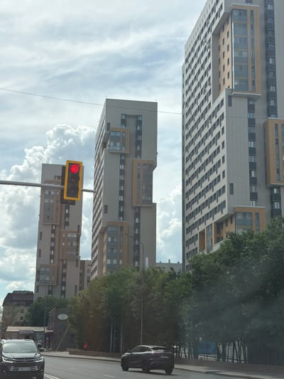This media file depicts a street view in Astana, Kazakhstan, featuring a modern high-rise building with a light yellow facade and numerous windows. The building's architecture is characterized by stacked sections, some with protruding balconies. In the foreground, a traffic light is visible, displaying a red signal. The sky is filled with dramatic white clouds against a pale blue backdrop, suggesting daytime with variable weather. To the right of the building, lush green trees line the street, with a darker building and more trees visible further in the distance. Two cars are present on the road: a black car in the middle distance and a black car in the foreground, with a partial view of its license plate, "707 ST 750". The scene appears to be an urban environment, possibly a residential or commercial district, with a sense of everyday activity implied by the traffic signal and vehicles. The overall impression is one of a contemporary cityscape on a bright, cloudy day.