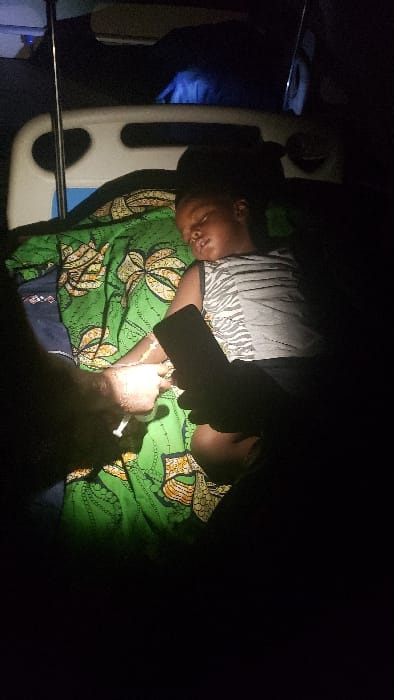 The scene depicts a medical intervention in Narayi, Nigeria, occurring in low light conditions. A young, dark-skinned child, approximately 5-10 years old, lies on a medical bed, covered by a green patterned blanket, appearing to be asleep or unconscious. The child wears a grey and white striped tank top. An intravenous line is inserted into the child's left forearm, secured with white tape. A light-skinned adult hand on the left is holding a syringe, connected to this IV line, actively administering a substance. Another hand, dark-skinned, is visible on the right, holding a black smartphone, with its screen illuminated, potentially providing additional light to the immediate area. A white hospital bed frame and an IV pole are partially visible in the dimly lit background.