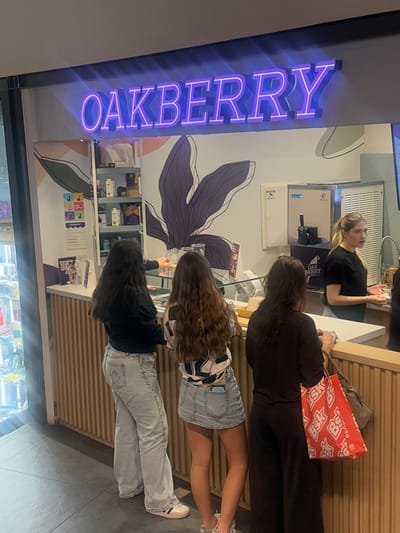Three female patrons stand at the counter of an "Oakberry" store. A female employee is behind the counter. The storefront is identified by a vertically-oriented, neon purple "Oakberry" sign. Inside, there is shelving with products for sale and a decorative wall mural of a dark purple flower. One patron has a red "BSK" bag. This scene is in the city of Matosinhos, Portugal.