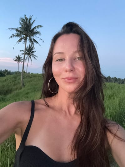 The photograph depicts a woman in a black top, wearing a gold hoop earring. She is positioned in front of a green field dotted with palm trees under a dusk sky. The background suggests a tropical location, consistent with areas in General Luna, Philippines, where palm trees and open fields are typical features. The woman is facing the camera, with a slight smile. The photograph appears to have been taken at ground level, possibly as a selfie.