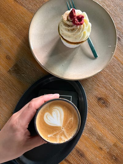 A top-down shot captures a well-lit, slightly angled view of a wooden table, showcasing a cappuccino with a heart-shaped design in the foam held by a hand. The cappuccino sits on a small black tray. To the left is a dessert on a plate, possibly a cupcake, adorned with white frosting and a garnish of dried red fruit, accompanied by a small fork. The image has no clear indicators that could confirm it was taken in Meisenheim, Germany. No people other than the visible hand are discernible, and the environment is a simple indoor cafe setting.