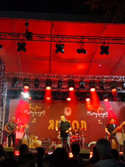 The image depicts an outdoor concert stage illuminated by red lights. On the stage, there are several musicians performing. In the foreground left, a man in a grey t-shirt and dark pants plays an electric guitar. To his right, a lead singer in a black t-shirt and blue jeans holds a microphone and sings. Behind them, a drummer in a black t-shirt is seated at a drum kit. On the far right, a man in a black t-shirt and grey pants plays a bass guitar. A large banner hangs behind the musicians with text that appears to be in Cyrillic script, reading "Бири Манджаро" which translates to "Beer Stew" or "Beer Goulash". Above this text, a logo for "Ямбол" (Yambol) is visible, indicating the concert is taking place in the city of Yambol, Bulgaria. The stage is equipped with professional lighting rigs, speakers, and other musical equipment. A crowd of spectators is partially visible in the foreground, their faces blurred due to the low light and distance. The overall atmosphere suggests a lively evening event.