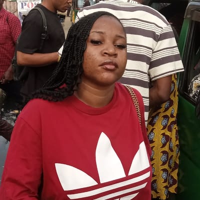 A young woman with long braids is wearing a red Adidas shirt with a gold chain strap visible on her shoulder. She looks directly at the camera with a slight pout. The background is a busy street scene with pedestrians, and a vehicle interior with striped and patterned upholstery. The angle suggests the photo was taken while she was lying down or in a reclining position.