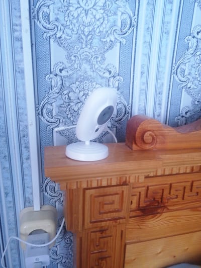 The image shows a close-up of a baby monitor on a wooden shelf next to a wall with patterned wallpaper. The baby monitor is white and has a round base. The wallpaper is blue and white with a damask-like pattern. To the right of the monitor, there is a wooden headboard with intricate carvings. A white electrical cord is visible, plugged into a cream-colored wall socket. The lighting suggests it is daytime. The scene is likely a bedroom or nursery. The image does not show any people or animals.