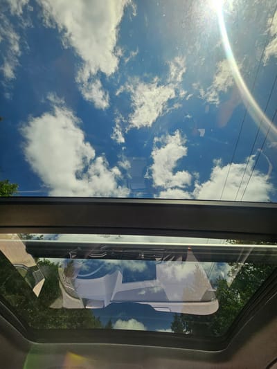 The image is a view looking up through an open car sunroof on a sunny day. The sky is blue with scattered white clouds. The sun is visible in the upper right corner, causing a lens flare. Below the sky, the reflections in the sunroof show a white car, trees, and some power lines. The scene appears to be in a residential area or a park, given the presence of trees. The specific location in Issaquah, United States of America, cannot be determined from the image alone.