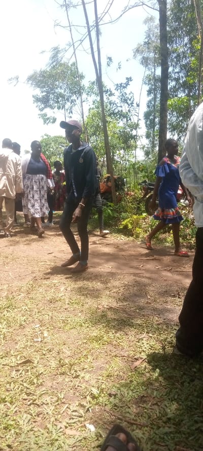 The image shows a group of people standing and walking in a wooded area in Malava, Kenya, located at 0.508 latitude and 34.791 longitude. The people are dressed in casual attire, and there are a few motorcycles parked nearby. The ground is covered in dirt and leaves. The scene appears to be a typical outdoor gathering in a rural setting in Kenya.  A man in a black jacket and black cap is walking towards the right side of the image. He appears to be the subject of the photo.  There is a young woman wearing a blue dress standing nearby.  Another woman wearing a floral dress is walking behind the man.