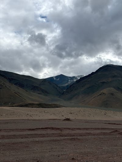 The image depicts a vast, mountainous landscape under a cloudy sky. The foreground is dominated by a rough, gravelly terrain with patches of darker soil. In the middle ground, a lighter, sandy slope rises, dotted with small rocks and some mounds of earth. The background features a range of imposing, dark green mountains with rugged slopes, some of which are partially obscured by mist or clouds. The sky is filled with heavy, grey clouds, with some lighter patches suggesting the presence of the sun trying to break through. There are no people or animals visible in the scene. The overall impression is one of a remote and stark, natural environment. This image is of the natural landscape surrounding Nyoma, India.