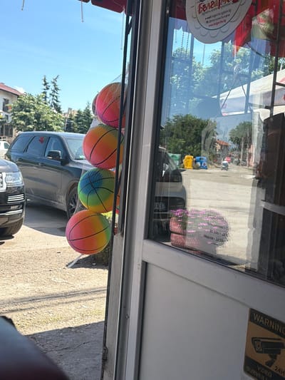 A low-angle, outdoor, eye-level shot shows the exterior of a small shop in the city of Ravno Pole, Bulgaria, on a sunny day. In the foreground, three multi-colored toy balls are hanging in a net, with the bottom two balls partially obscured by the edge of the frame. The balls are rainbow-colored, with sections of pink, orange, yellow, blue, and green. To the left, a dark-colored SUV is parked. The shop has a large glass window, through which several reflections are visible, including trees, buildings, and a street scene with yellow and blue bins, and a person on a scooter. A sign with Bulgarian text is visible on the window, stating "РАБОТНО ВРЕМЕ" (Working Hours). A yellow and black "WARNING VIDEO SURVEILLANCE" sticker with a camera icon is affixed to the bottom right of the shop's exterior. The ground is paved with gravel and concrete.