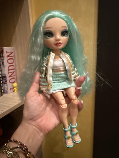 The image shows a close-up of a doll with light blue hair and striking blue eyes, held in a person's hand. The doll is wearing a textured light blue and gold striped jacket over a white top and a matching light blue skirt. Its feet are adorned with light blue sandals. The doll's fingers have long, white nail polish. The person's hand is supporting the doll, and their wrist is decorated with several bracelets, including a braided one with red, white, and blue threads, and a silver chain bracelet.
In the background, on a shelf-like surface, there is a small item with the word "LONDON" printed in red, stylized letters, with an image of a train beneath it. The lighting suggests an indoor setting, likely with artificial light, and the background is blurred, focusing attention on the doll. The overall impression is a casual, personal photograph, possibly a snapshot of a toy or collection. The presence of the "LONDON" item provides a subtle hint of travel or interest in the city.
