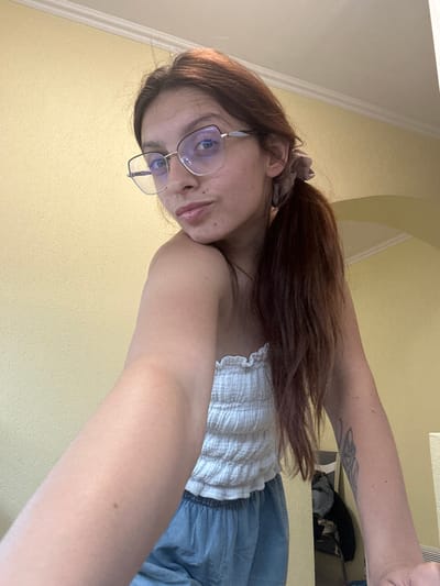 A young woman with auburn hair pulled into a ponytail and wearing glasses is taking a selfie in what appears to be an indoor setting. She is wearing a white, strapless, smocked top and light blue pants. Her arm is extended forward, and a portion of her arm is visible in the foreground, showing a tattoo on her left forearm. The background is a textured, pale yellow wall, and a white crown molding runs along the top of the wall. The lighting suggests an indoor environment, possibly a room in a residential building. The image does not contain any information that would allow for the identification of its location within the city of Tryavna, Bulgaria.