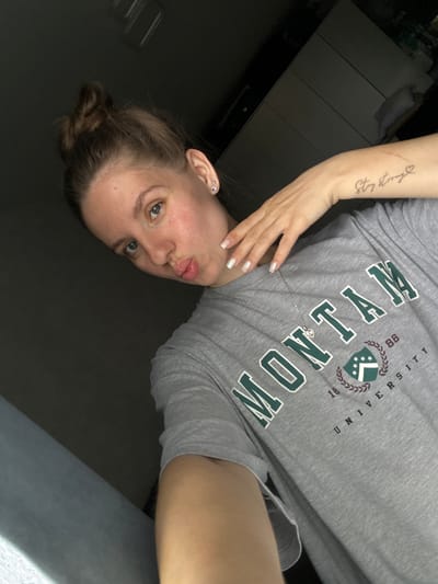 A woman with fair skin, blue eyes, and light brown hair pulled back in a bun is positioned in the lower left of the frame. She is looking at the camera with a slight pouting expression. She wears a grey T-shirt with the words "MONTANA UNIVERSITY 1888" printed in green. A silver necklace is visible on her chest. The woman's left arm is raised with her hand near her neck. A tattoo is visible on her forearm. To the left of her is a dark, glossy surface that could be a table or counter. A light grey object appears at the bottom right corner. The location, Klin, Russia, cannot be determined from this image.