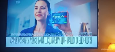 The image shows a close-up of a television screen displaying a pharmaceutical advertisement. In the center, a woman is holding a blue box of "НАЛГЕЗИН" tablets. The background is blurred, showing a bright, possibly indoor environment. Below the woman and the product, a large, bold text reads "САМОЛІКУВАННЯ МОЖЕ БУТИ ШКІДЛИВИМ ДЛЯ ВАШОГО ЗДОРОВ'Я". There is also smaller, disclaimer-like text below that. In the bottom right corner of the screen, a timestamp "01.07.2026 00:11" is visible. The image quality suggests it is a photograph taken of a screen, possibly in a home or retail setting. There is no specific information about the city of Odesa, Ukraine within the image content.