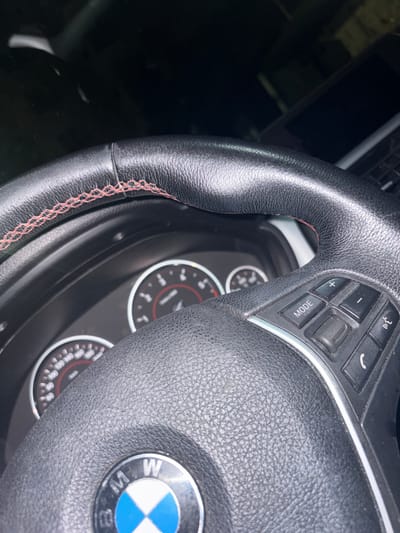 The image shows a close-up view of a car's steering wheel and dashboard. The steering wheel is black leather with red stitching and features several buttons, including "MODE," volume controls (+/-), and call/end buttons. The BMW logo is visible on the steering wheel's center. The dashboard includes a speedometer and tachometer, with red and white markings. A portion of a laptop is visible in the background. The lighting suggests it is nighttime or a dimly lit environment. The image was taken inside a car, likely parked. There is no visible indication of the city of Dupnitsa, Bulgaria, or any specific location within it.