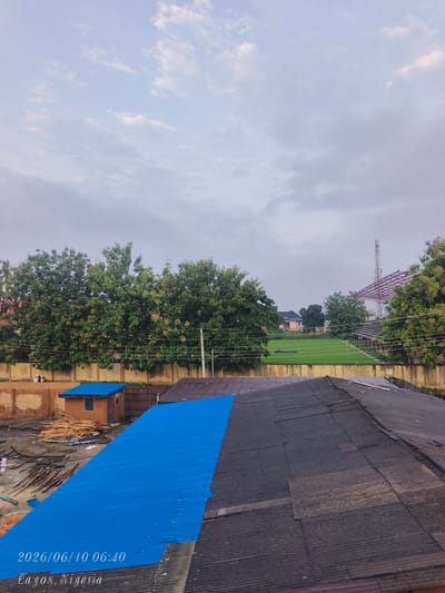 The image depicts an aerial view of a construction site in Shangisha, Nigeria, on June 10, 2026, at 06:40. In the foreground, two sections of rooftops are visible: one with a bright blue corrugated metal roof and another with a darker, weathered corrugated metal roof. Piles of lumber and construction materials are scattered on the ground near a small, orange-brown structure with a blue roof. In the midground, a tall concrete wall separates the construction area from a green field, possibly a sports field, with stadium seating and a transmission tower in the distance. Lush green trees line the perimeter of the field. The sky is overcast with scattered clouds, suggesting early morning or a cloudy day. Two indistinct figures are visible on top of the concrete wall in the distance.