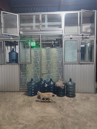 In Sagoe, Indonesia, a ginger cat walks across a concrete floor in what appears to be a storage area. The cat is the primary subject in the foreground. In the background, there is a large shelving unit made of aluminum and glass. Several large blue water jugs are visible on the floor in front of the shelving unit. One of the jugs has a cloth draped over it. The shelving unit has multiple compartments, some of which are open and contain the water jugs. One compartment has a green light and some electronic equipment. The walls are made of a corrugated metal material. The overall environment appears to be indoors and dimly lit.
