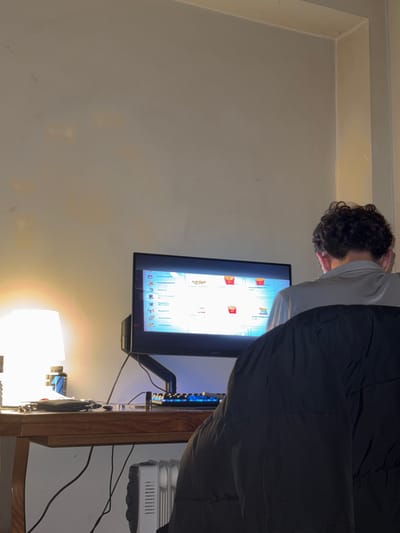 An indoor scene captures a person engaging with a computer monitor, likely in an evening or nighttime setting. The image is oriented vertically but depicts a person with dark, curly hair, seen from behind and the side, leaning slightly forward as they face a brightly lit screen. They are wearing a light-colored top and dark clothing.
The setting appears to be a room, possibly a bedroom or home office, with plain, light-colored walls. A wooden desk or table is positioned beneath the monitor. On the desk, a lamp emits a warm, bright glow, providing a significant light source in the otherwise dimly lit room. To the right of the monitor, a white electronic device with visible vents and controls, resembling a humidifier or small air purifier, sits on the desk. Numerous cables are strewn across the desk surface, connecting the various electronic items.
The computer monitor displays a graphical user interface, which appears to be a digital menu or ordering system, possibly from a fast-food establishment. Several food icons are visible, including what look like french fries in red containers and various beverages. Prominently displayed text on the screen, in Portuguese, includes categories such as "Menu Infantil" (Children's Menu), "Complementos" (Complements), "Pratos" (Dishes), "Bebidas" (Drinks), "Acompanhamentos" (Accompaniments), and "Sobremesas" (Desserts). The overall impression is one of someone browsing or selecting items from a digital menu.