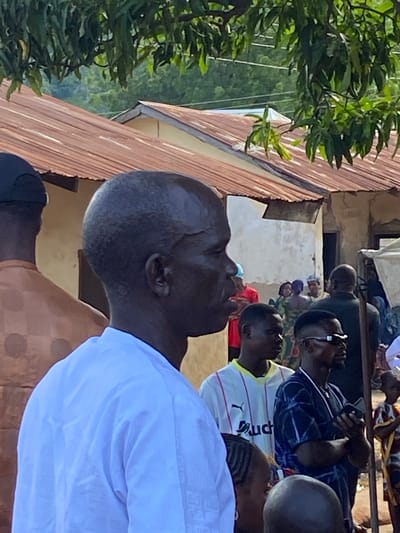 This image depicts a community gathering in Mararaba, Nigeria, under bright daylight. In the foreground, a dark-complexioned man in profile faces right, wearing a white patterned tunic with a raised collar. To his immediate left, the back of another individual wearing a brown patterned garment and a dark cap is visible. Further right, in the midground, are several other individuals: a young man in a white and black striped PUMA-branded shirt with "Auchan" text looks left; next to him, a man in a dark blue patterned shirt and dark sunglasses holds a mobile phone, looking right. Behind these individuals, a group of people, including women in colorful attire and head coverings, are visible. To the far right, a partial view of a child in patterned clothing is present. In the background, multiple single-story buildings with rusty corrugated metal roofs and light-colored walls are visible. Overhead and in the distance, lush green foliage and trees indicate dense vegetation, with hills or further dense greenery visible under a clear sky. Wires, possibly power lines, traverse the sky above the roofs.
